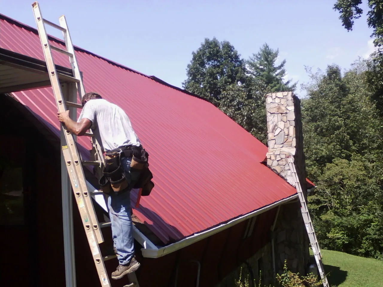 Roofer on a ladder beside a red metal roof and stone chimney — Soffit & Fascia Repair by Roofing Brothers in Lingle